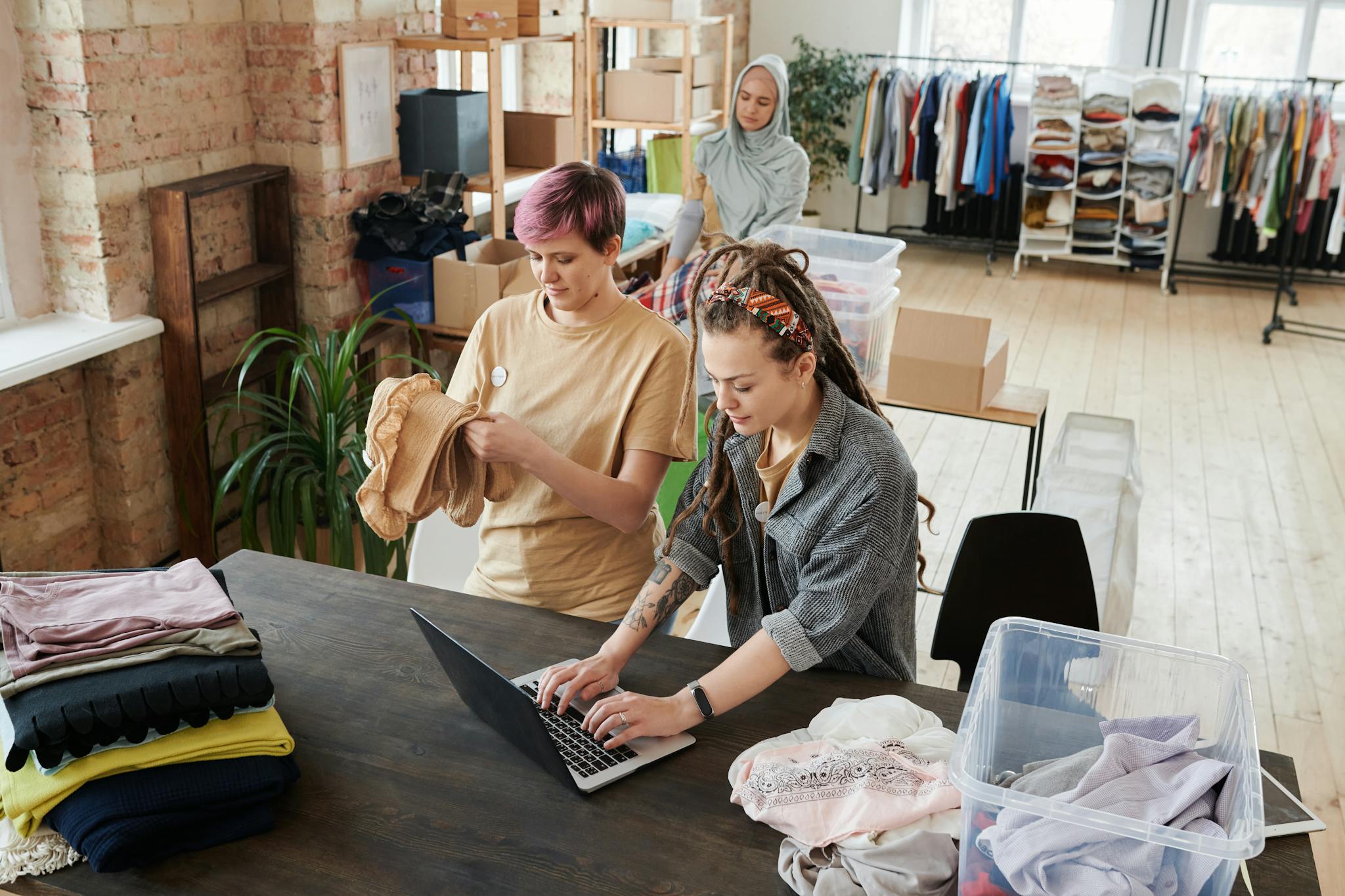 Women organizing clothes and using a laptop in a donation center. Diverse teamwork and collaboration.